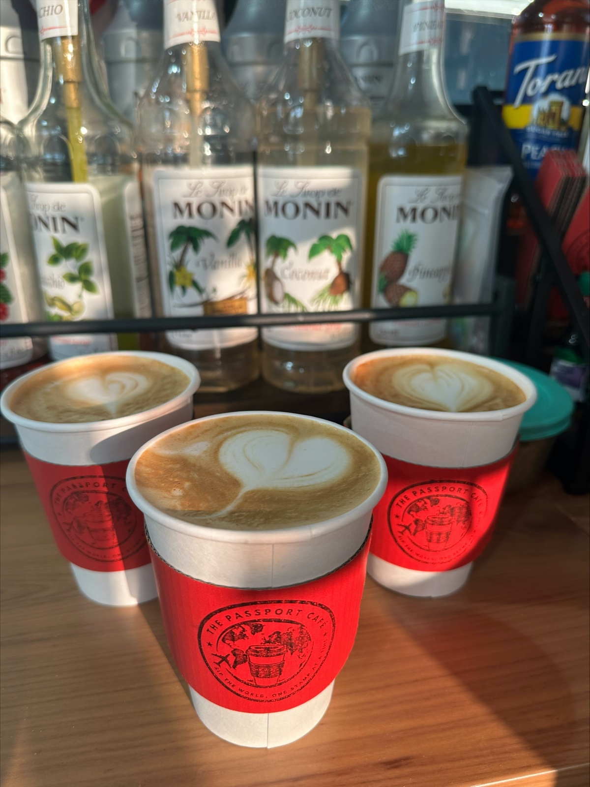 Three lattes with heart latte art in red stamped Passport Cafe sleeves